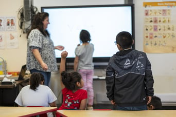 The image shows a classroom scene with a teacher and several young students. The teacher, standing on the left, is interacting with the students, some of whom are seated while others are standing near a large interactive whiteboard. One student is writing on the board, which displays the word "vest." Another student is seated with their hand raised, indicating they may have a question or want to participate. The classroom has various educational posters on the walls, including one with basic keywords and another with illustrations. The students are engaged in the lesson, and the atmosphere appears to be focused and interactive.