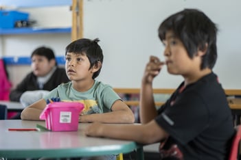 This image depicts a classroom setting with three young students seated at a table. The focus is on two boys in the foreground, with one boy wearing a light green shirt and looking attentively towards the front of the classroom. In front of him is a pink container filled with pencils and other writing tools. The other boy, who is slightly out of focus, is wearing a black shirt and appears to be deep in thought, with his hand near his mouth. In the background, another student is partially visible, also paying attention to the classroom activities. The atmosphere seems to be one of concentration and engagement.