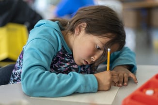 This image features a young girl intensely focused on her writing. She is leaning over a piece of lined paper, holding a pencil, and appears to be deeply engaged in her task. The girl is wearing a turquoise hoodie with a patterned design on the sleeves. Her concentration is evident from her posture and facial expression. The background shows a slightly blurred classroom setting with other students and classroom materials, suggesting a busy and active learning environment. The overall atmosphere is one of focus and dedication to schoolwork.